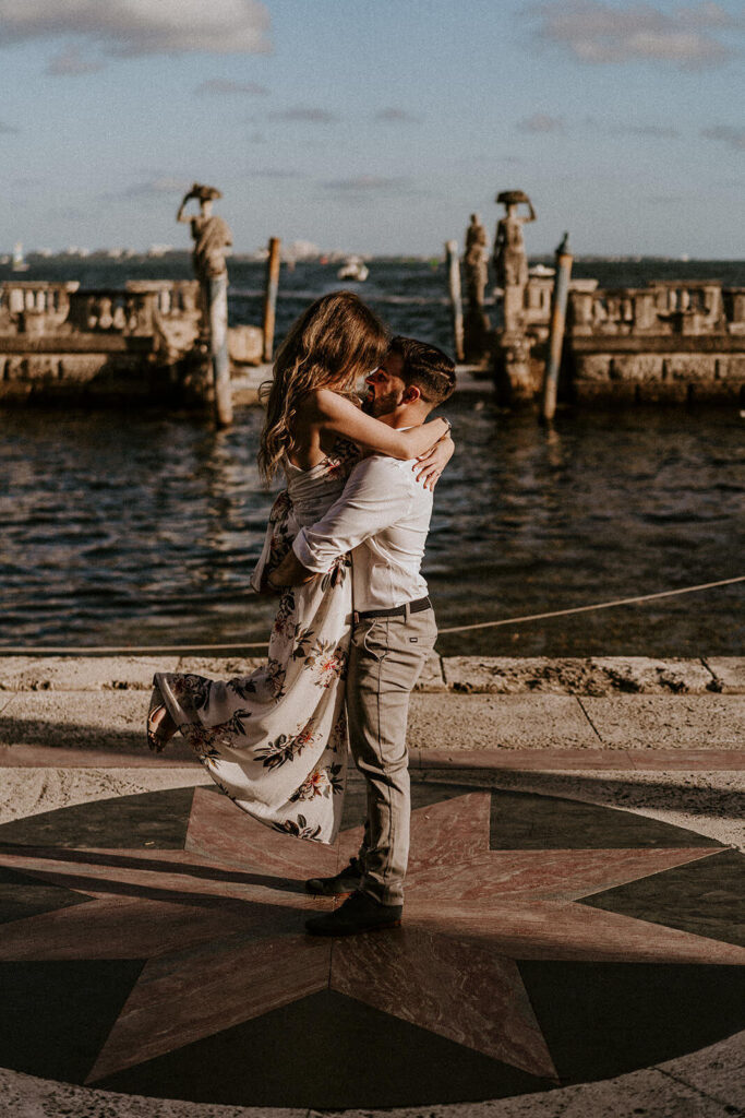 a man holding his fiance up in front of a water structure at their engagement shoot