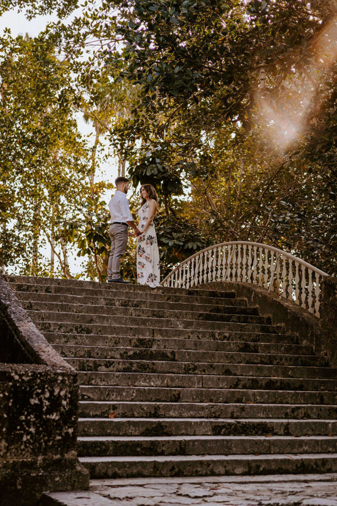 a couple stands at the top of a staircase with sunlight beaming down