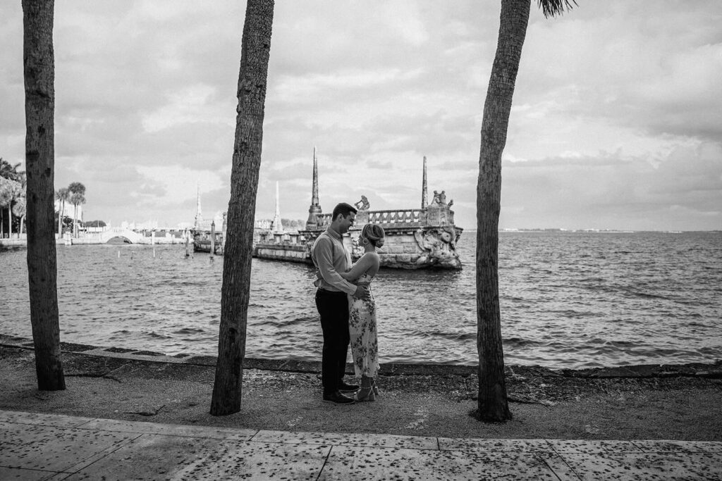 a couple stands between 2 palm trees in front of the water structure at Vizcaya Museum and Gardens