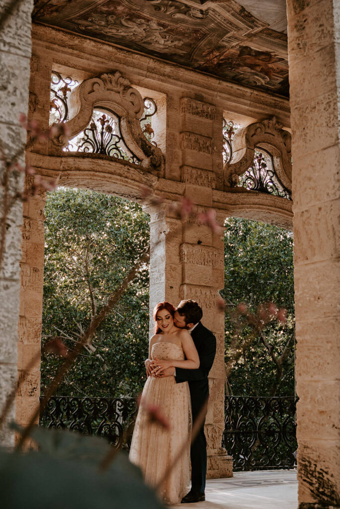 a groom hugs his bride from behind while displaying the beautiful ceiling in the building 