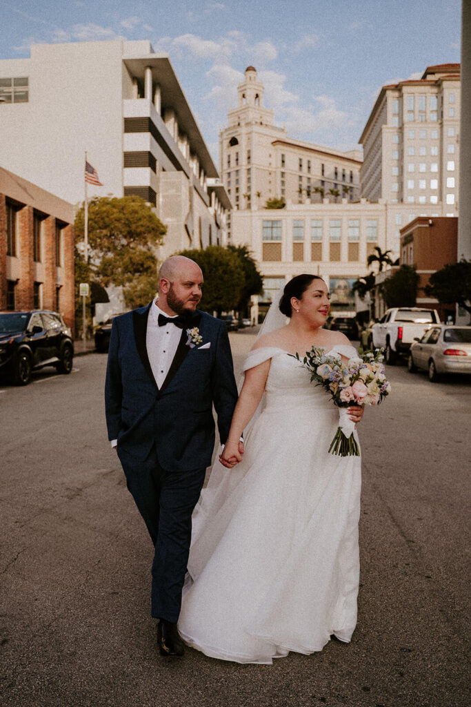 a bride and groom hold hands while walking down the street with the famous Loews hotel behind them in Miami florida