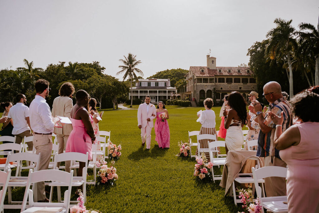 a bride and groom walk into their ceremony space hand in hand while their guests watch at their miami wedding