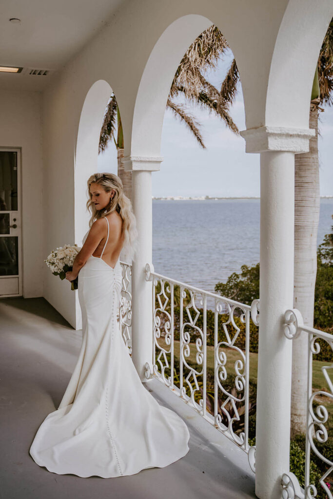 a bride poses with her back to the camera with her bouquet on a balcony overlooking the ocean