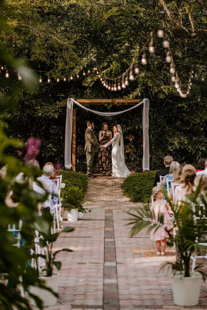 a bride and groom holds hands at their ceremony space under lights and surrounded by greenery