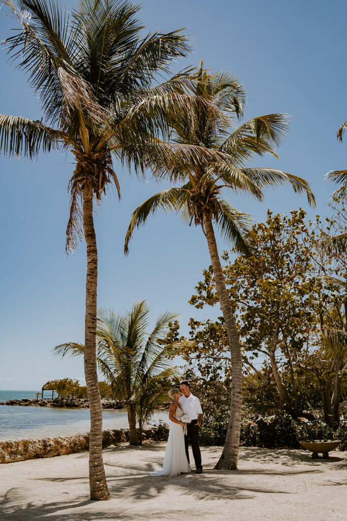 a couple poses under giant palm trees in front of the ocean at their spring time wedding