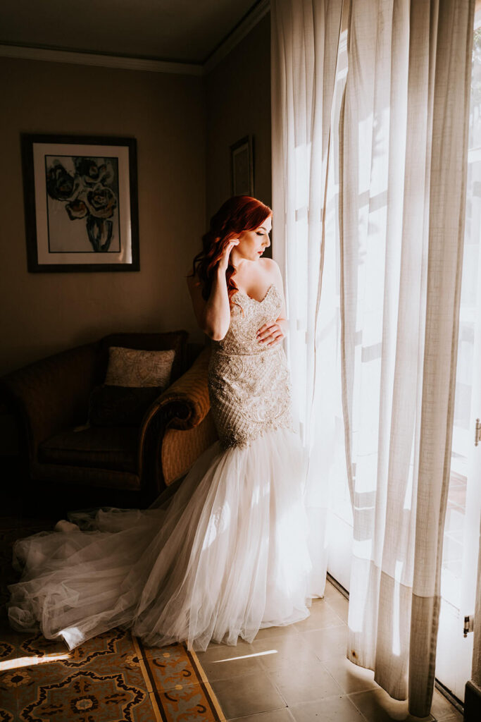 a bride with red hair gets ready for her wedding day by putting on her earring in front of the window at Villa Woodbine in Coconut Grove