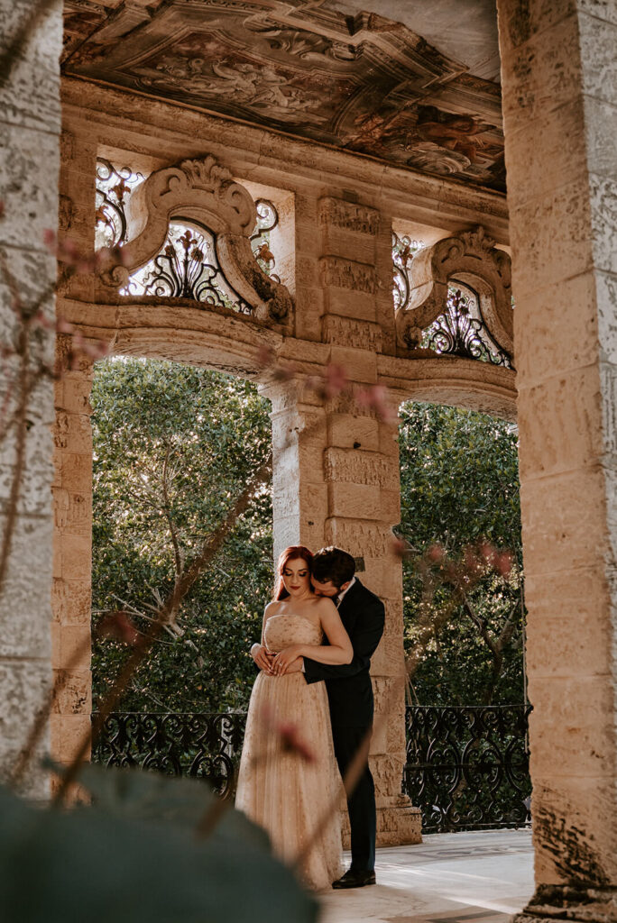 groom hugs his bride from behind in a structure located in the gardens of Vizcaya in Miami