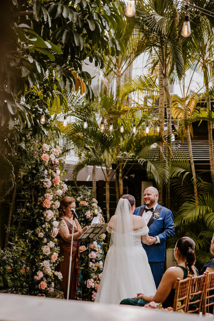a groom laughs at his wedding ceremony in Miami surrounded by florals and palm trees