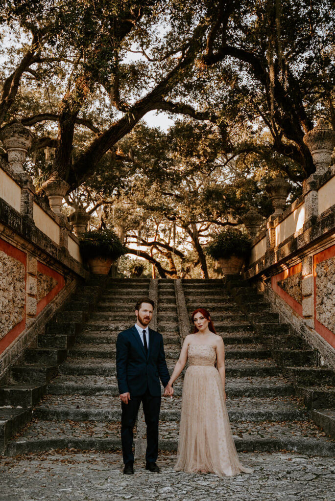 a couple holds hands and poses in front of a staircase in Miami at Vizcaya Museum