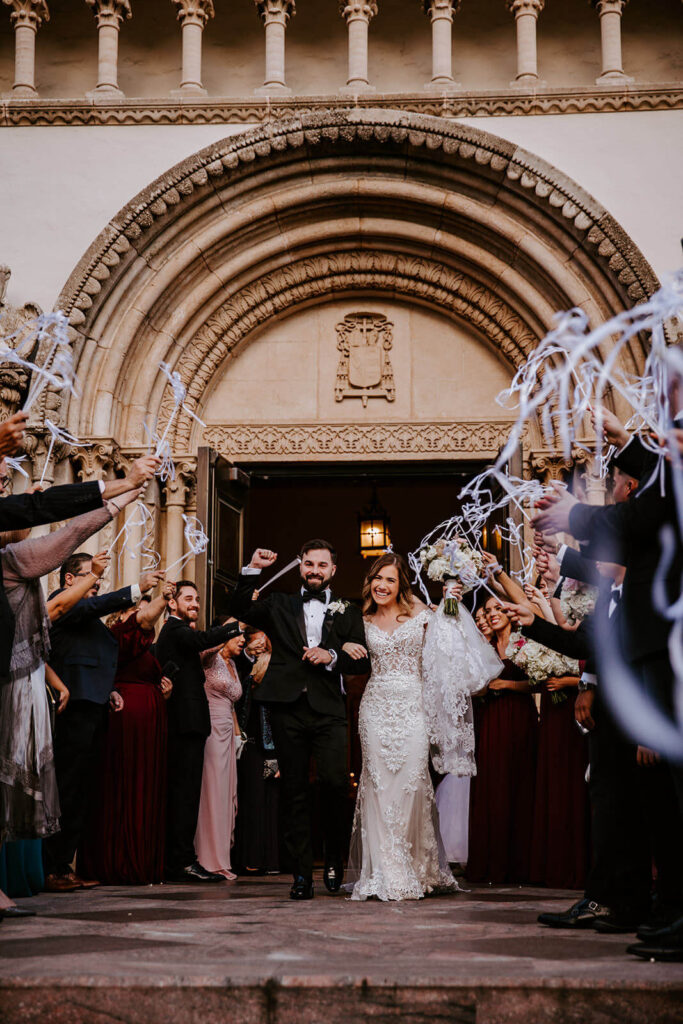 a bride and groom walks out of their miami wedding venue while their guests wave streamers
