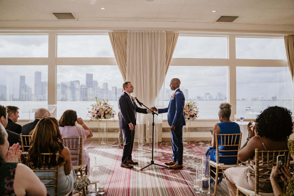2 grooms hold hands at their ceremony in front of Miami Beach with the ocean behind them