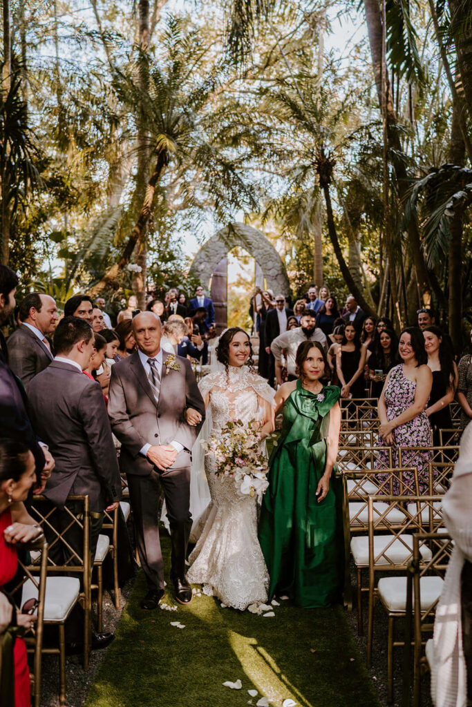 a bride walks down with aisle with her mom and dad surrounded by trees at her miami wedding