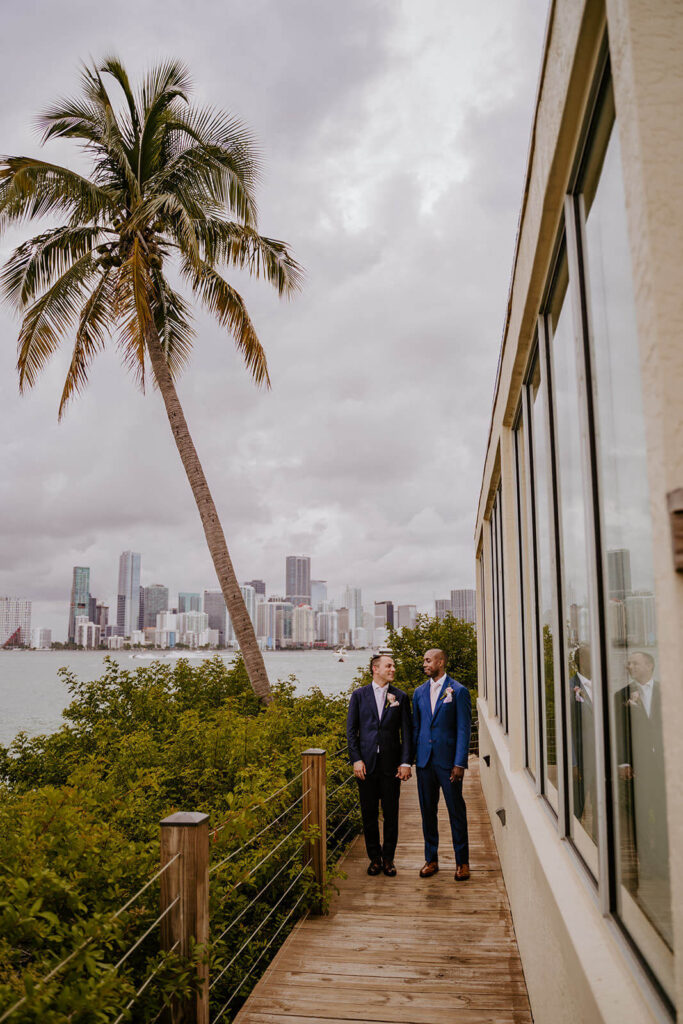 2 grooms hold hands while they walk with the miami skyline behind them at the Rusty Pelican