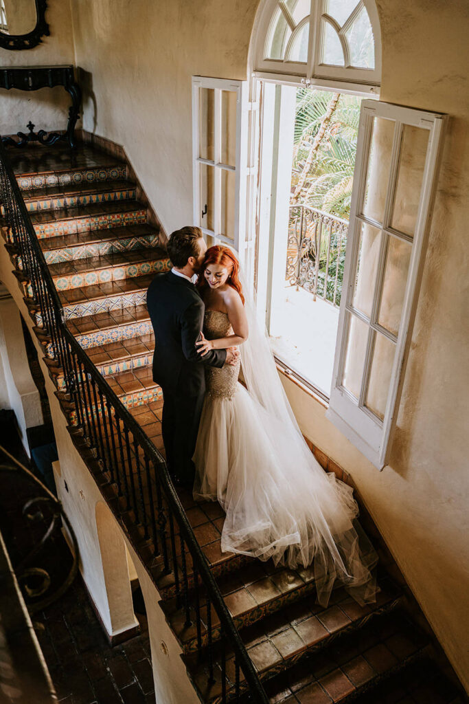 a groom kisses his bride on the cheek on the staircase at their wedding venue