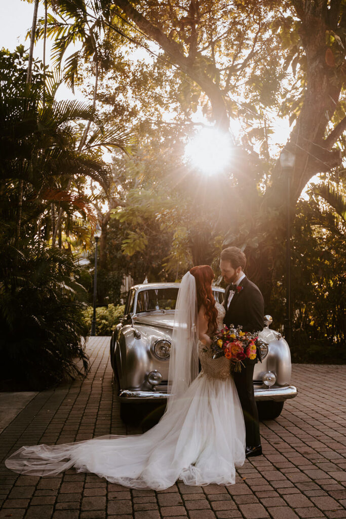 a couple kisses in front of a luxury car while the sunset is behind them