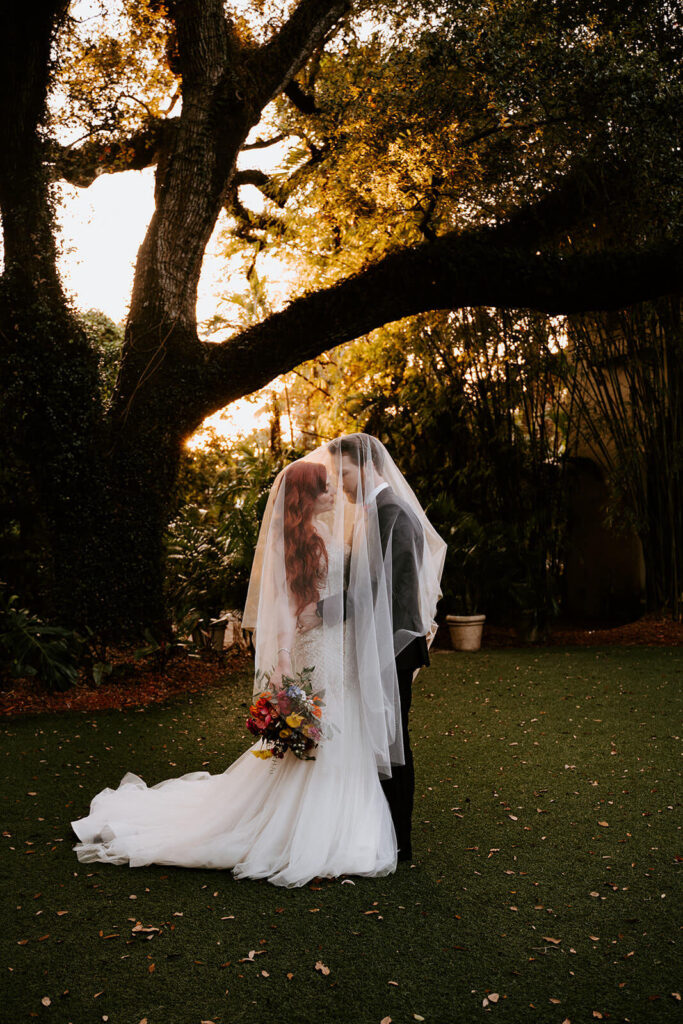 a couple kisses under the veil in front of the tree at Villa Woodbine