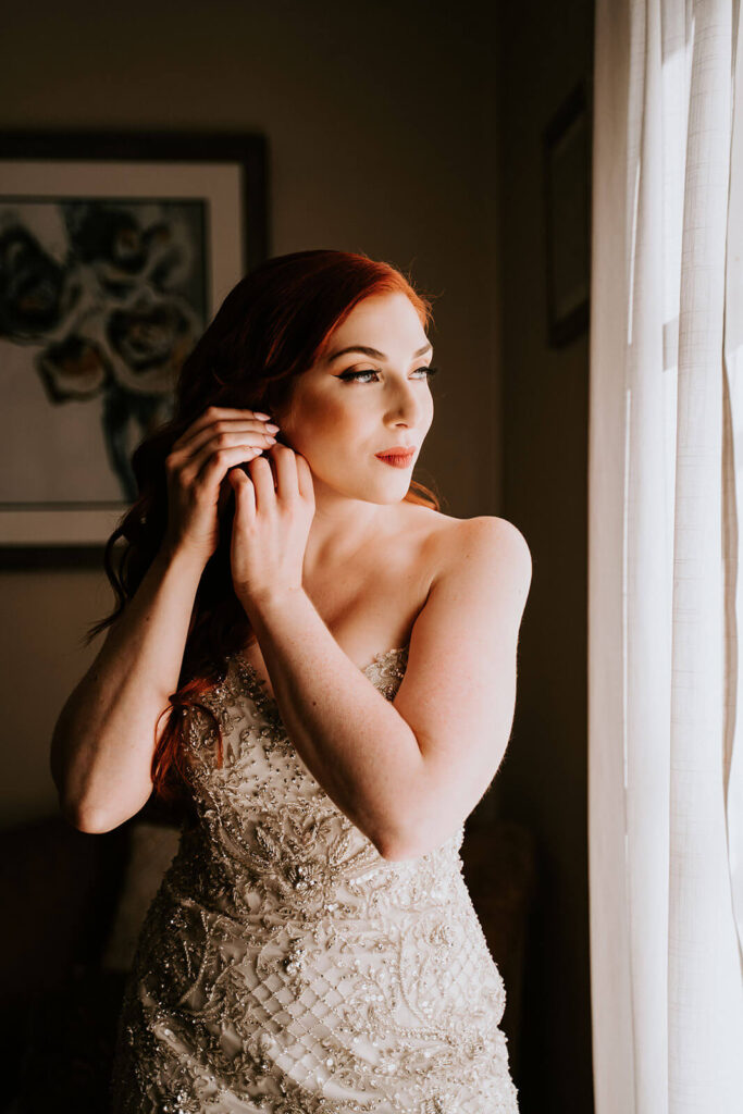 a closeup of a bride putting on her earrings while looking out of the windo